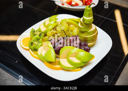 Vassoio di frutta ben organizzato con mele verdi, pere, uva, kiwi e arancia a fette, servita su un piatto bianco. Foto Stock