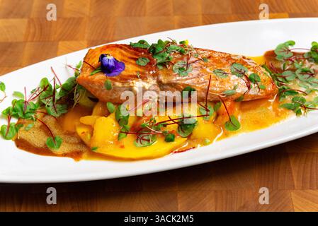 Piatto gourmet placcato con salmone scottato in padella servito su fette di mango con microverdure e una ricca salsa dorata, raffinato stile da pranzo. Foto Stock