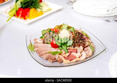 Elegante piatto di salumi a fette servito con lattuga, erbe aromatiche, pomodoro ciliegino, sottaceti e rafano su un tavolo bianco. Foto Stock
