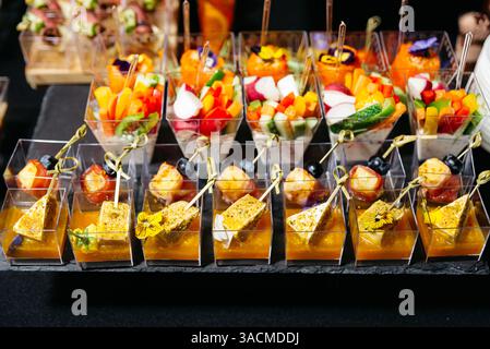Una varietà di eleganti antipasti serviti in bicchieri di plastica trasparente, con verdure, formaggi, frutta e fiori commestibili per eventi. Foto Stock
