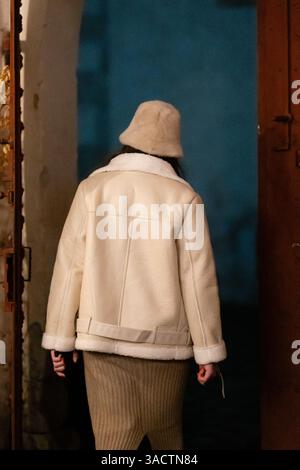 Un individuo elegante con un caldo cappotto bianco e un cappello beige si erge su una porta di legno, catturando un momento di solitudine alla moda in un ambiente con luce soffusa Foto Stock