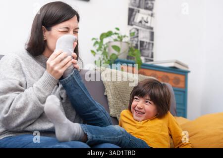 Un bambino gioioso ride mentre sua madre si toglie giocosamente i calzini. Un momento divertente e spensierato per tutta la famiglia in un ambiente domestico naturale. Foto Stock