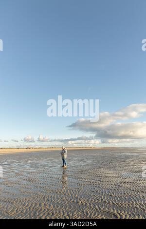 Una donna in abiti caldi si trova da sola nel mezzo del Mare di Wadden e guarda il suo cellulare con la bassa marea sulla spiaggia, Lakolk su Rømø, Den Foto Stock