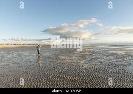 Una donna in abiti caldi si trova da sola nel mezzo del Mare di Wadden e guarda il suo cellulare con la bassa marea sulla spiaggia, Lakolk su Rømø, Den Foto Stock