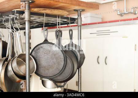 Appendiabiti con padelle e pentole in cucina. Assortimento di padella in ghisa, piastra e pentole in acciaio inox appese all'isola della cucina. Spazio Foto Stock