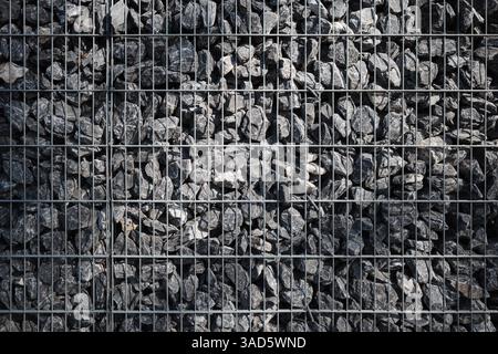 Una parete in gabbione molto compatta, costituita da rocce grigie grezze tenute insieme in una griglia metallica strutturata. Foto Stock