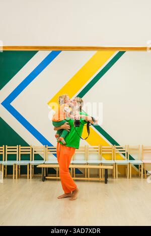 Un uomo tiene un bambino tra le braccia e il bambino indossa una camicia verde. La scena è ambientata in una stanza con molte sedie, e l'uomo è in piedi Foto Stock