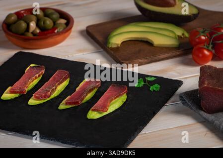 Piatto di pietra con fette di avocado ricoperte di fette di mojama, accompagnato da olive, avocado e pomodori ciliegini, una tipica tapa dell'Andalusia. Foto Stock