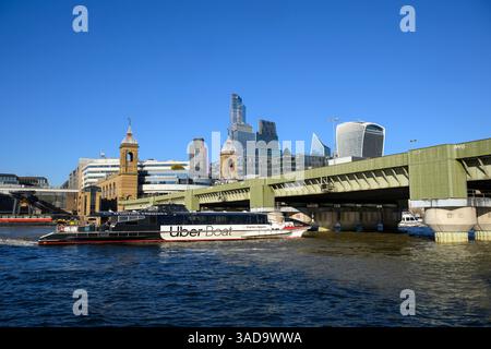 Un Uber Thames Clipper passa sotto il ponte ferroviario e la stazione di Cannon Street, con i grattacieli del quartiere finanziario della City of London in t Foto Stock