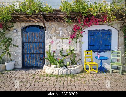 Terrazza rustica e mediterranea. Pareti in pietra adornate da fiori e vegetazione vibranti, porta e finestra in legno blu, sedie colorate e tavolo aggiunti Foto Stock