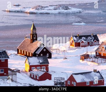 Una pittoresca scena invernale caratterizzata da un piccolo villaggio con case colorate in legno e una chiesa, circondata da neve e ghiaccio. Il paesaggio comprende un fr Foto Stock