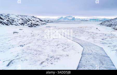 Un tranquillo paesaggio invernale caratterizzato da un percorso tortuoso attraverso un terreno innevato. La scena include colline rocciose e montagne lontane sotto un cielo nuvoloso Foto Stock