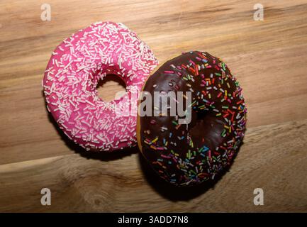 Due deliziose ciambelle si trovano su una tavola di legno, una ricoperta di glassa rosa e spruzzi bianchi, e l'altra con una ricca ricopertura di cioccolato e una vivace m Foto Stock