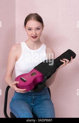La giovane e attraente musicista femminile sta togliendo l'ukulele dalla custodia nera morbida e guardando la fotocamera. Allegra giovane donna adulta musicista in abiti casual Foto Stock