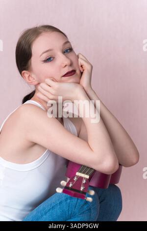 Annoiata musicista donna che riposa la testa sulle mani mentre sta in piedi sull'ukulele. Bella giovane donna che guarda la macchina fotografica con un'espressione calma. Esecutore che riposa du Foto Stock