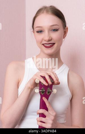 Ritratta giovane musicista felice con ukulele, il suo volto è pieno di gioia e guarda la macchina fotografica con un sorriso sincero. Bella donna positiva Foto Stock