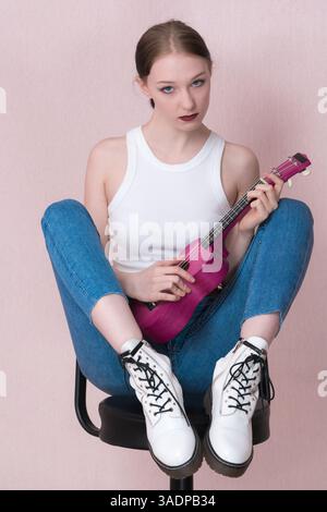La giovane musicista suona ukulele, siede sullo sgabello da bar e guarda direttamente nella fotocamera con un'espressione vuota. Una bella donna indossa un abbigliamento casual Foto Stock