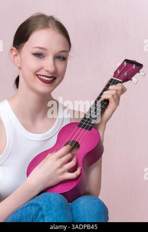 La giovane musicista sorride mentre suona ukulele, guardando la telecamera. La bella donna caucasica con gli occhi blu ama suonare la musica e il suo entusiasmo Foto Stock