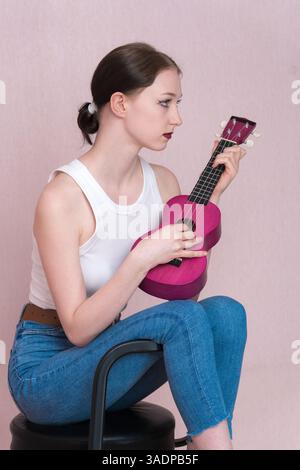 Giovane musicista donna seria che suona ukulele, siede su sgabello da bar e guarda lontano mentre succhia strumento musicale. Vista profilo donna sottile in Foto Stock