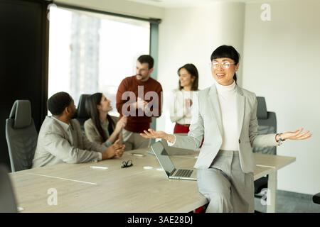 Gruppo di professionisti diversi impegnati in vivaci discussioni in un ufficio illuminato dal sole. Una donna è in piedi con fiducia, mostrando lavoro di squadra e collaborazione Foto Stock