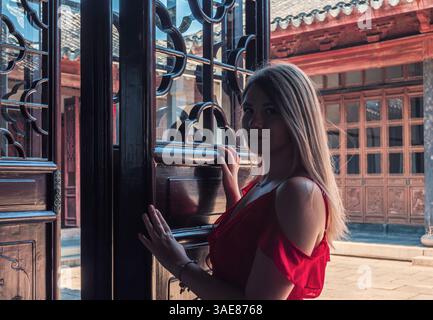 Una giovane donna con un vestito rosso si erge su una porta di legno ornata. Foto Stock