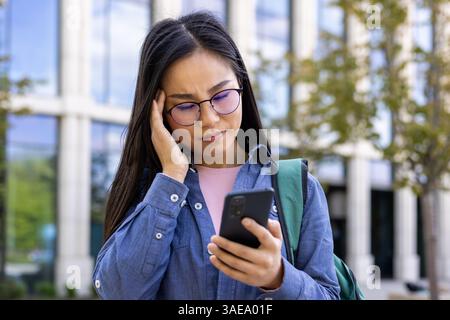 Una donna asiatica che indossa gli occhiali sta guardando il suo telefono con un'espressione preoccupata. Sembra stressata, forse a causa di un messaggio. Foto Stock