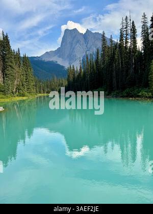 Un tranquillo lago turchese riflette gli alberi sempreverdi circostanti e una montagna torreggiante sotto un cielo blu parzialmente nuvoloso. Foto Stock