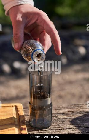 Processo di macinatura del caffè: Vista ravvicinata di una mano che versa i chicchi tostati in un macinacaffè da viaggio, all'aperto, in una mattinata di sole. Foto Stock