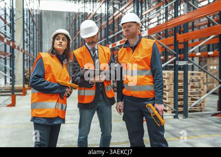 I lavoratori del magazzino e i responsabili che indossano giubbotti di sicurezza e cappelli rigidi controllano l'inventario utilizzando scanner di codici a barre e appunti in una grande distribuzione c Foto Stock