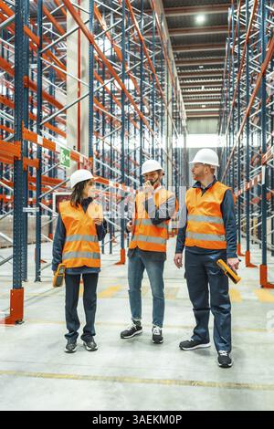 I lavoratori del magazzino che indossano giubbotti di sicurezza e caschi stanno discutendo di logistica, utilizzando walkie talkie e scanner di codici a barre, in un grande centro di distribuzione Foto Stock