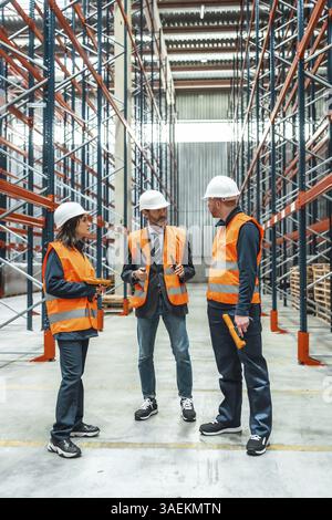 I lavoratori del magazzino e i responsabili che indossano giubbotti di sicurezza e cappelli di protezione discutono della logistica all'interno di un centro di distribuzione Foto Stock