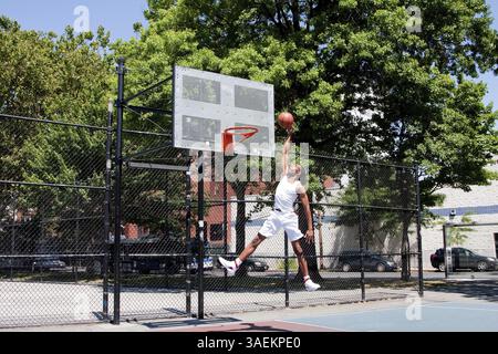 Sporty bello afro-americano di uomo vestito di bianco di saltare in aria per raggiungere il cesto mentre si gioca a basket in un tribunale aperto su una somma Foto Stock