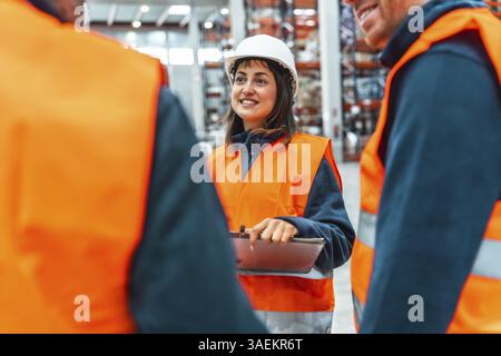 Gli addetti al magazzino che indossano giubbotti di sicurezza e cappelli rigidi discutono di logistica mentre utilizzano un tablet digitale in un magazzino di grandi dimensioni Foto Stock