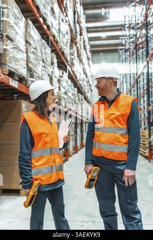 Due addetti al magazzino che indossano giubbotti di sicurezza e cappelli rigidi discutono di logistica, tenendo in mano scanner di codici a barre e un portadocumenti in una corsia di magazzino Foto Stock