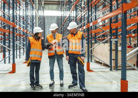 I lavoratori del magazzino che indossano giubbotti di sicurezza e cappelli rigidi controllano l'inventario utilizzando un tablet digitale e uno scanner per codici a barre in un grande centro di distribuzione Foto Stock