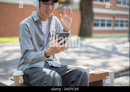 Un uomo asiatico sorridente con gli occhiali sta salutando il suo telefono, facendo una videochiamata o registrandosi, seduto sulla panchina del marciapiede. Passare il tempo libero. Foto Stock