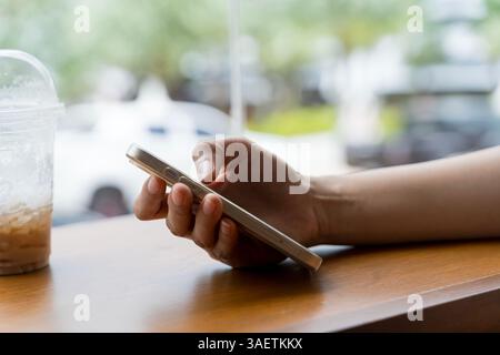 Mano di donna asiatica che usa lo smartphone su un tavolo di legno nel bar. Immagine ravvicinata di un uomo che tiene in mano e usa uno smartphone con una tazza di caffè su una tazza di legno Foto Stock