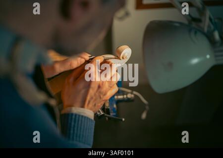Luthier intaglia meticolosamente un pezzo di legno, fissato in una morsa, usando una goffa, sotto il caldo bagliore di una lampada, mettendo in mostra la maestria artigianale in cui è coinvolta Foto Stock