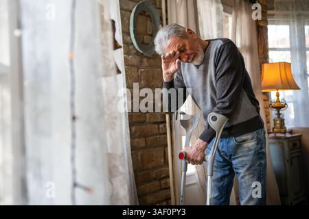 Vecchio con la stampella che si sente deluso, perso in pensieri tristi da solo a casa. Vedova uomo maturo. Foto Stock
