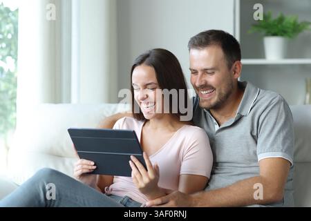 Coppia felice che controlla contenuti divertenti sul tablet seduto su un divano a casa Foto Stock