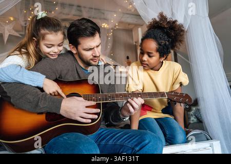 Felice padre single che insegna alle sue figlie e ai suoi figli multietnici e diversificati a suonare la chitarra a casa. Foto Stock