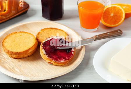 Spalmare la marmellata di mirtilli sul pane tostato con un coltello, accompagnato da succo d'arancia, burro e pane intrecciato, crea una scena di colazione sana e deliziosa Foto Stock