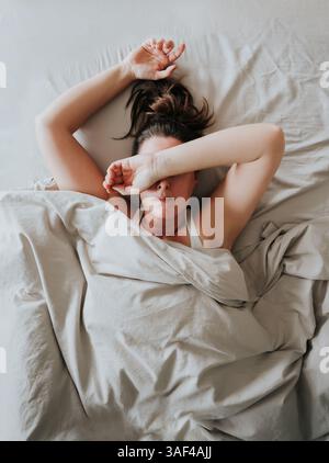 Vista dall'alto della donna stanca a letto con il braccio sopra la testa e gli occhi coperti. Foto Stock
