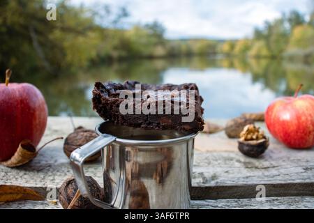 Picnic sul lago tranquillo. Tazza di metallo con caffè caldo o tee sul molo di legno sul tranquillo lago. Paesaggio naturale autunnale. Mele rosse, foglie autunnali, Foto Stock
