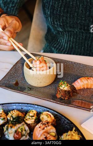 Una persona che usa le bacchette per raccogliere un pezzo di salmone da una piccola ciotola, con vari sushi e antipasti su un piatto rustico in primo piano Foto Stock