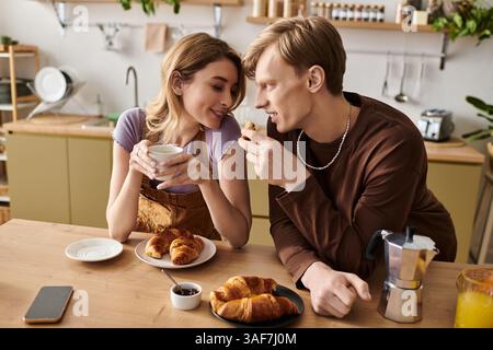 Una giovane coppia condivide con gioia la colazione con croissant e bevande calde nella loro luminosa cucina. Foto Stock