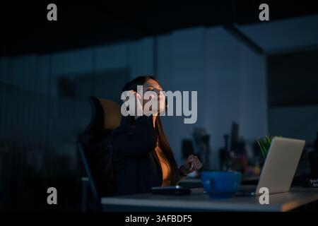 Donna sorridente con le cuffie che alzano le mani per festeggiare mentre si siede a una scrivania in un ufficio poco illuminato. Concetto di successo, gioia al lavoro, successo Foto Stock