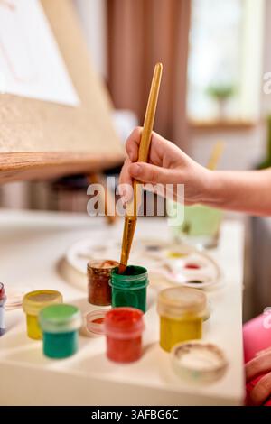 Primo piano verticale della mano del bambino che immerge il pennello nel vaso di pittura durante l'attività di apprendimento creativo a casa. Foto Stock