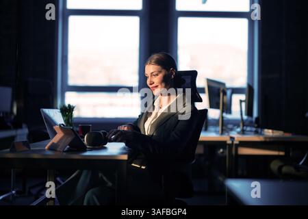 Professionista IT femminile o ingegnere IA che lavora fino a tardi in un ufficio moderno, illuminato dalla luce calda del sole. Concetto di tecnologia, sviluppo di software Foto Stock