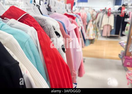 Una fila colorata di abiti per bambini è appesa ordinatamente a pennarelli bianchi in una corsia ben illuminata Foto Stock